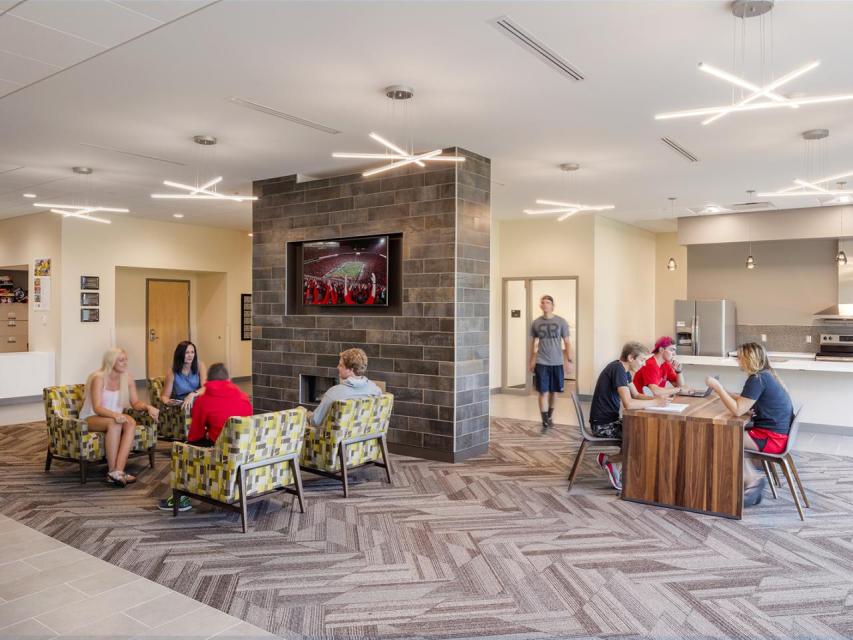 A group of students sits around a fireplace with a mounted tv while another group sits at a table near a kitchenette.