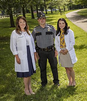 white female in lab coat standing smiling, man in officer uniform standing smiling, white female with black hair standing and smiling