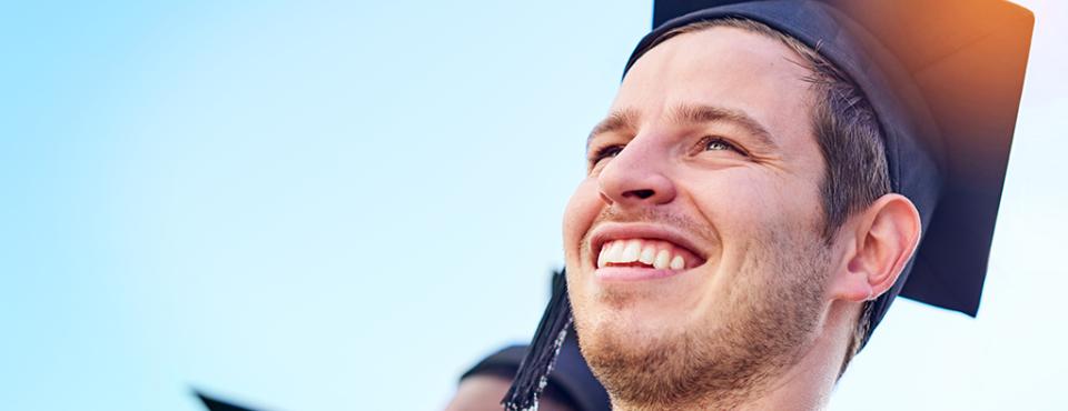 A male wearing a graduation cap and gown looks upward smiling. 