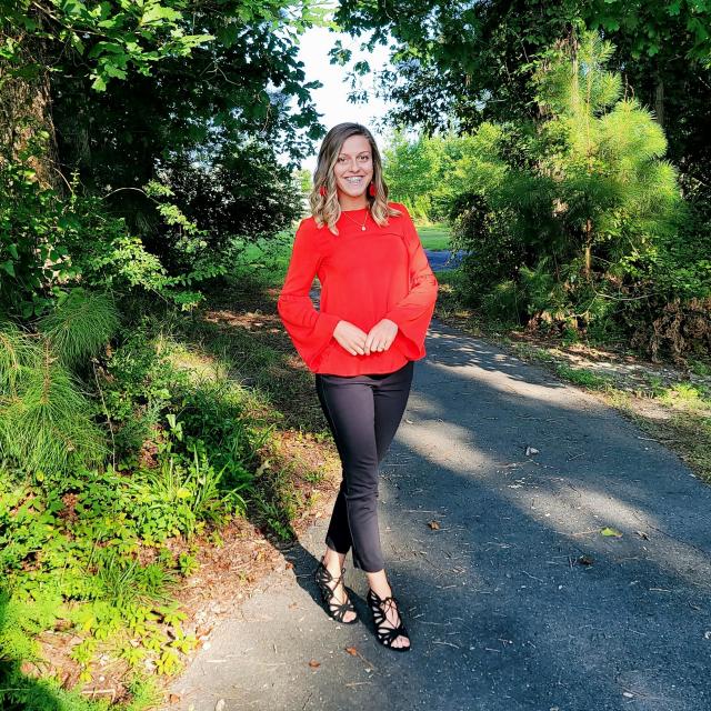 young Caucasian woman with blonde hair and blue eyes in red shirt, black pants, and black sandals poses for a picture