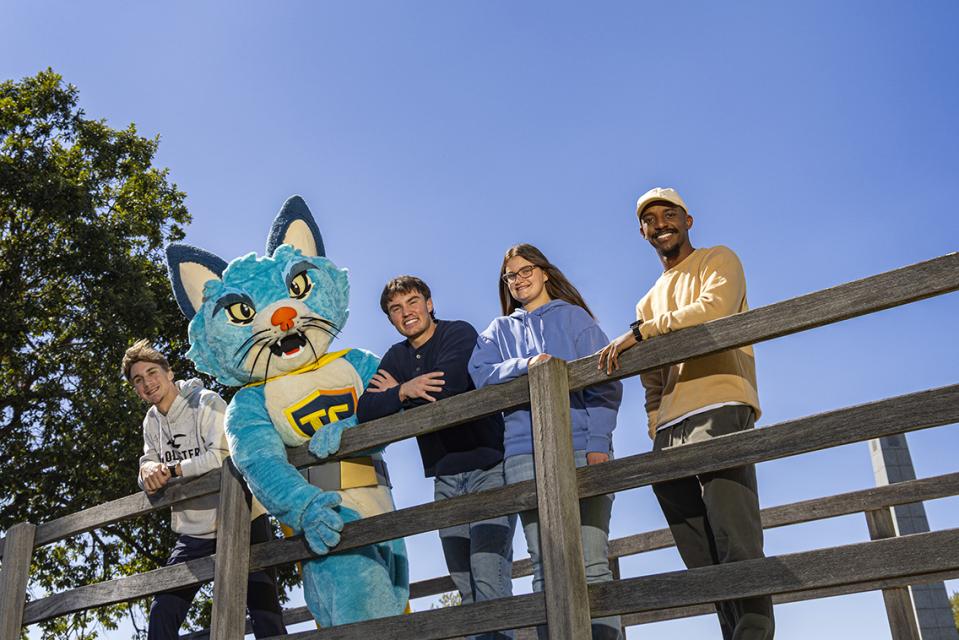 Four students stand on the iconic bridge on COTC's Newark campus with TC the mascot.