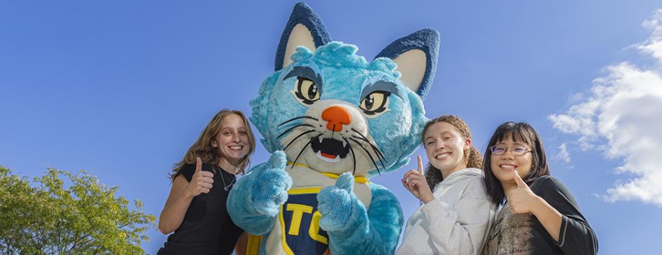TC the blue cat mascot gives two "thumbs up" as three female students surround him giving "thumbs up" gestures.