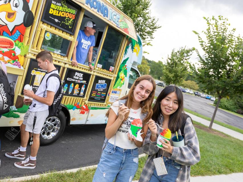 Two smiling female students stop for a portrait with their Kona Ice as other students line up at the truck behind them during Welcome Week.